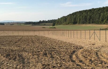 Ein weitläufiges Feld mit bearbeitetem Boden und einem Zaun im ländlichen Gebiet, umgeben von Wäldern und Hügeln.