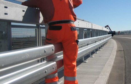 Ein Bauarbeiter in orangefarbener Arbeitskleidung führt Sicherheitsarbeiten an einer Brücke durch, während ein Bagger im Hintergrund zu sehen ist.