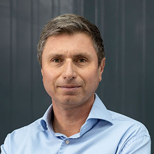 Porträt eines Mannes mit kurzen Haaren in einem hellblauen Hemd, vor einer neutralen grauen Wand.