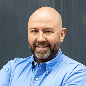 Ein Mann mit Glatze und Bart trägt ein blaues Hemd, lächelt freundlich und steht vor einem grauen Hintergrund.