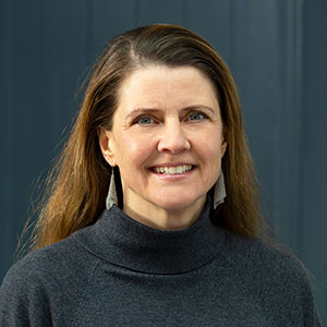 Porträt einer Frau mit langen, braunen Haaren, die in einem dunklen Pullover lächelt. Sie steht vor einer blauen Wand.