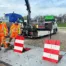 Drei Bauarbeiter in Warnkleidung arbeiten mit Verkehrsabsperrungen auf einer Baustelle, während ein Lkw im Hintergrund parkt.